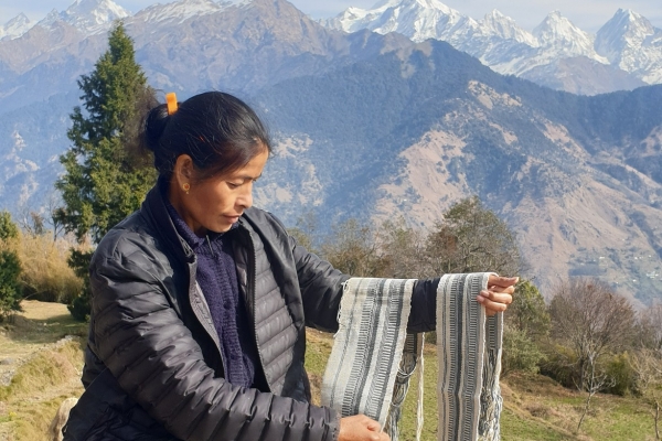 A woman sits holding up textiles woven on a traditional loom, against a backdrop of the snowcapped Himalayas