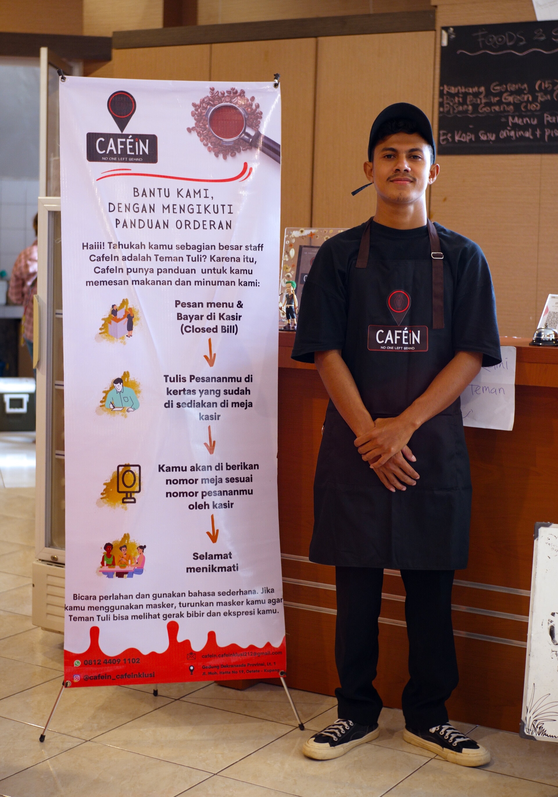 Barista Doni Kopong standing by a banner which advises customers at CafeIn: to order in writing at the cashier, pay up front, and take off their masks to allow deaf staff to lip read. 