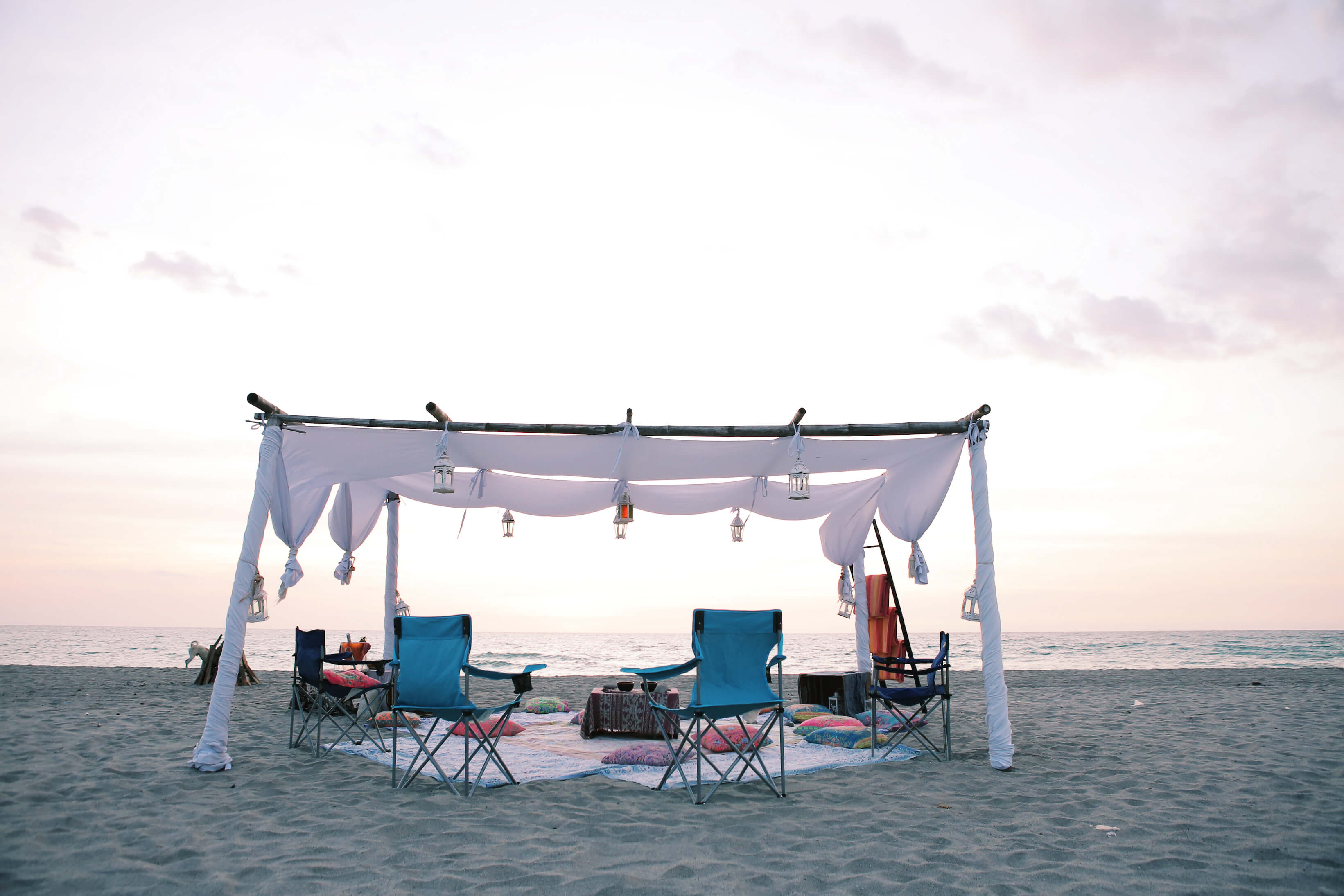 Camping chairs and beach blankets are arranged artfully beneath a gauzy tent with the setting sun in the distance.
