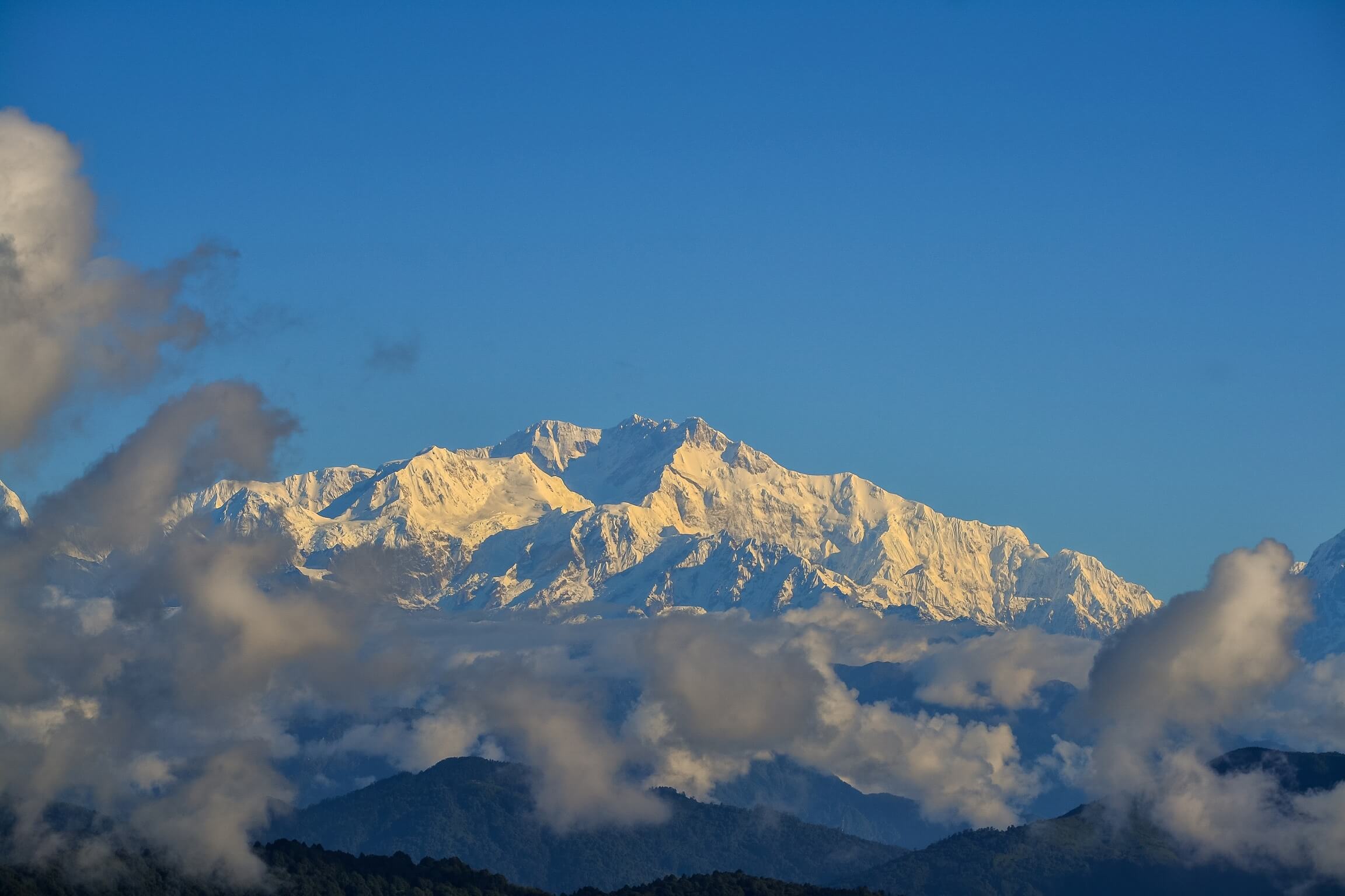A mountain swathed in snow against a bright blue cloudless sky, with light cloud cover in the foreground.