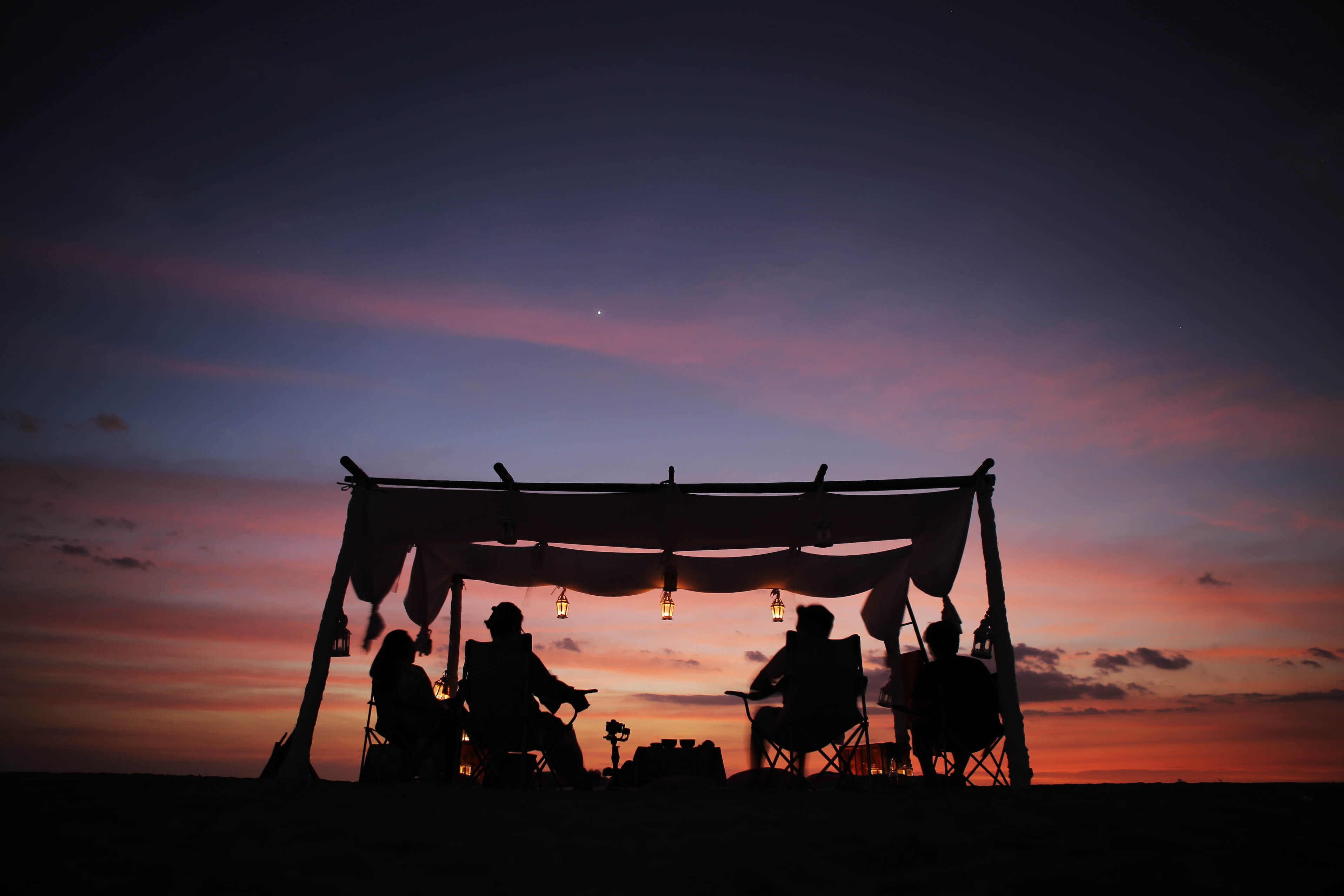 A group of people sit under tent watching a dazzling sunset in the background.