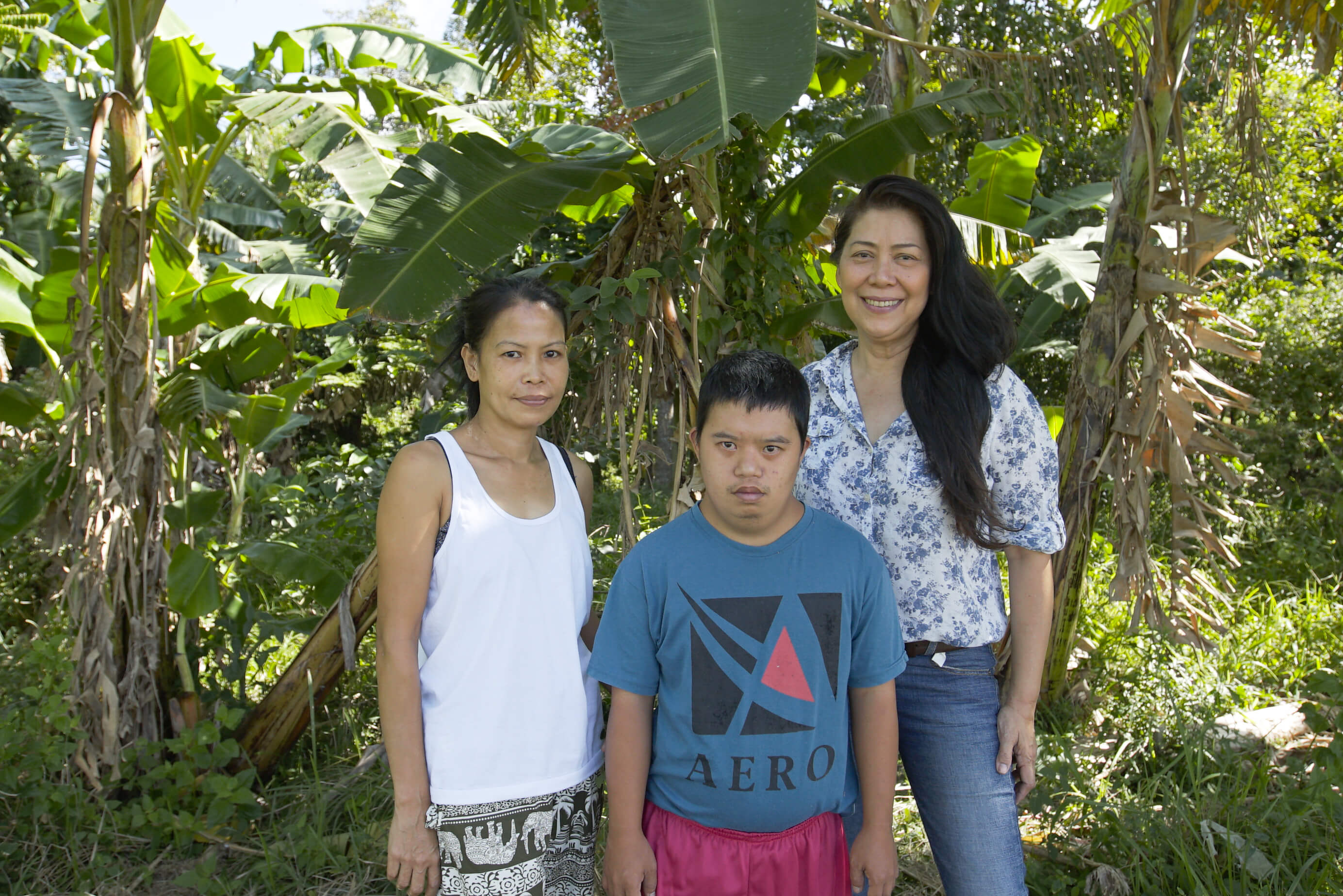 Two women stand with a man between them under the shade of a banana tree.