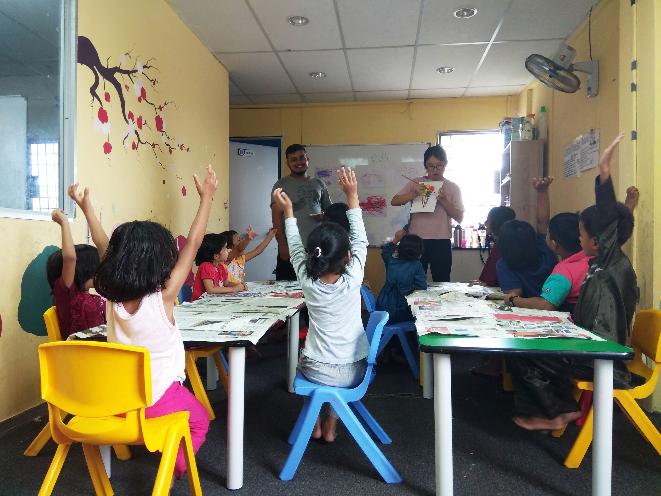 A group of children raise their hands in excitement in an art class at a refugee school in Malaysia supported by Advocates for Refugees - Singapore
