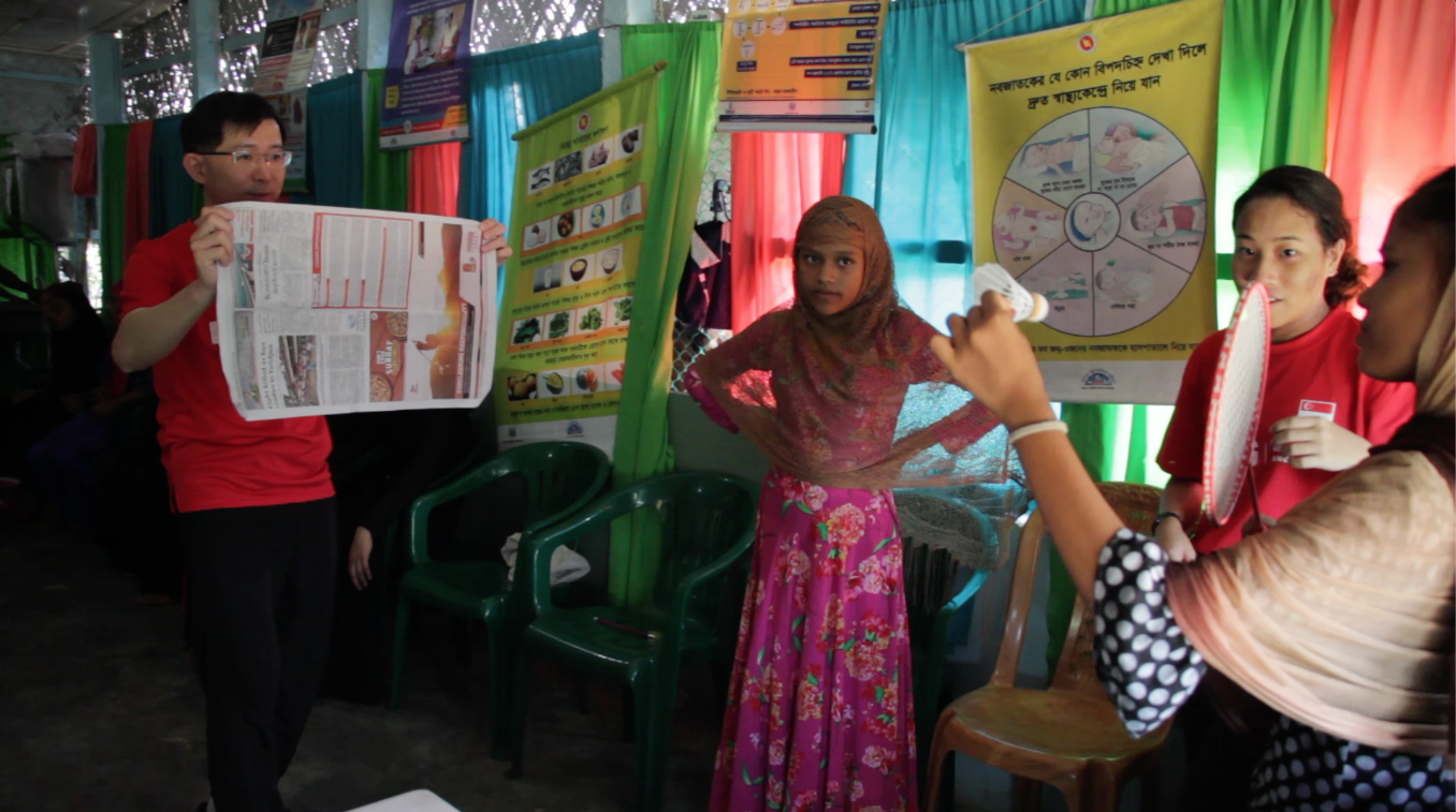 A refugee girl holds a shuttlecock in her hand as she readies to serve for a game of badminton, while a friend and a volunteer from Relief Singapore look on with encouragement on the side. 