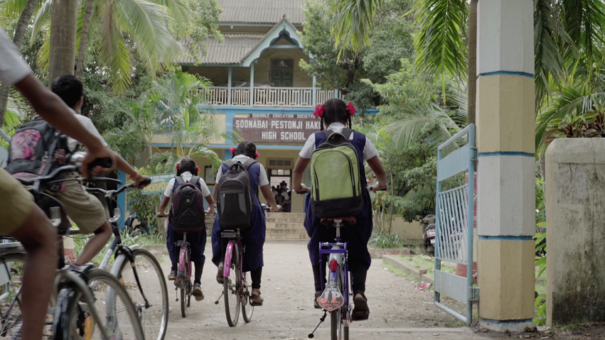 a group of children in school uniforms cycling through the gates into their school in the background.